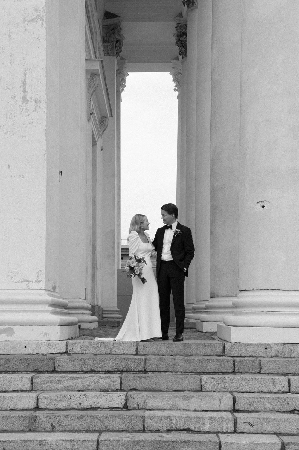 Wedding couple in formal attire posing between elegant white pillars.