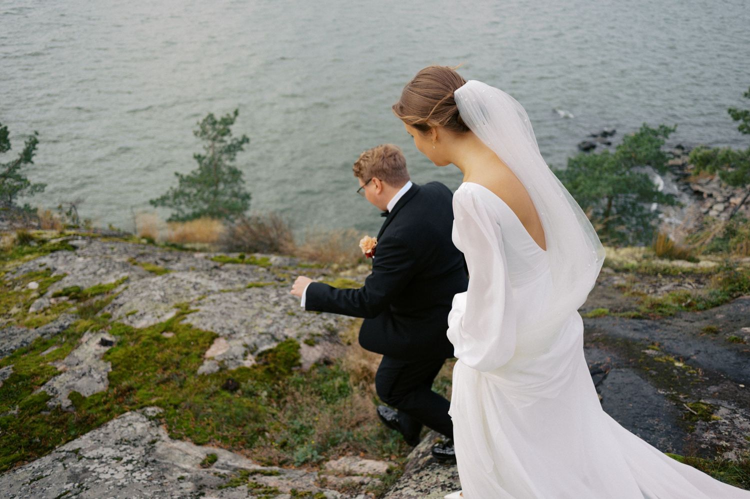A couple in wedding attire stands on a rocky cliff overlooking a serene lake with evergreen trees in the background.