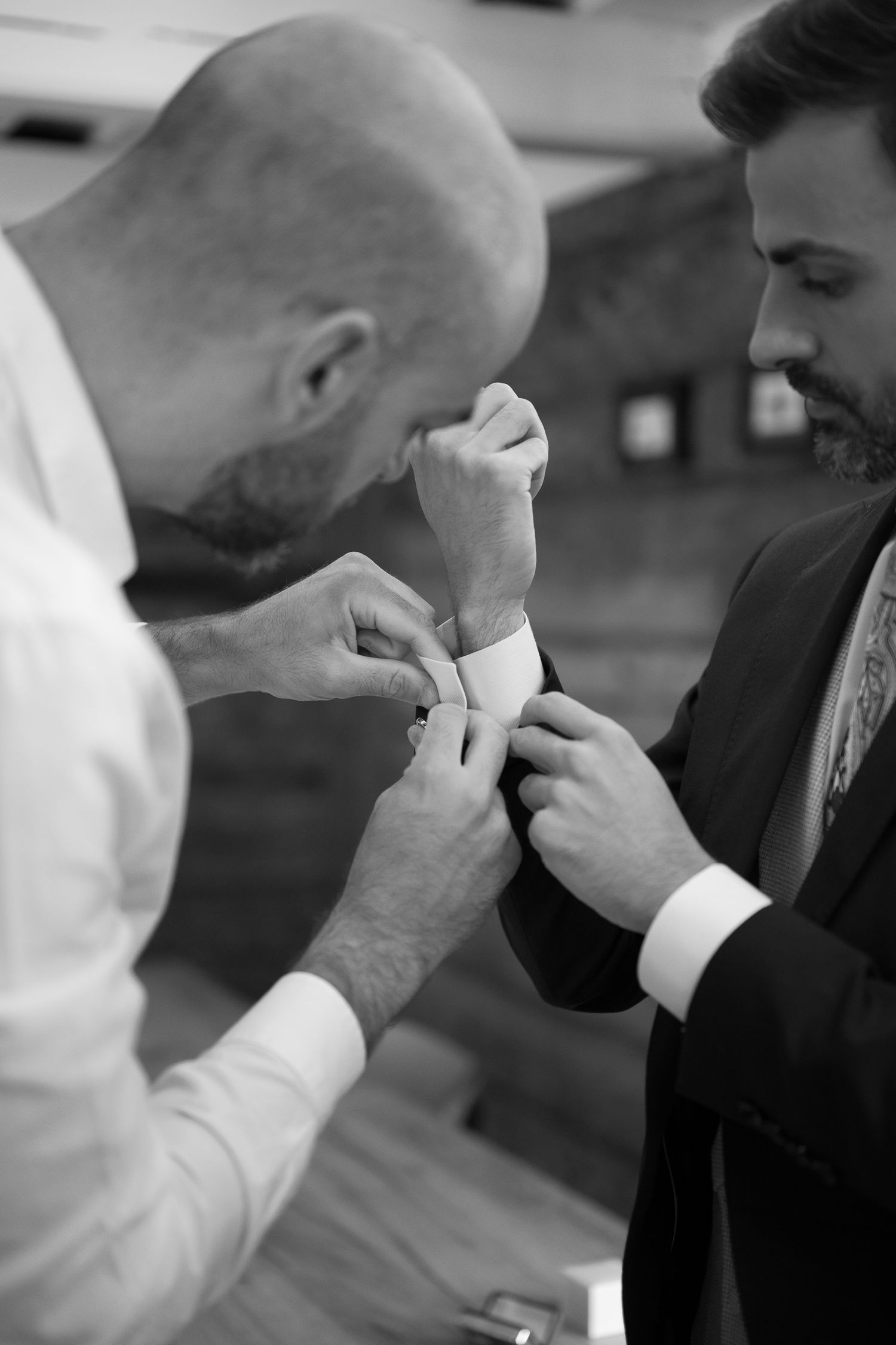 Black and white photo of a bald man helping a bearded groom fasten his cufflinks before a wedding ceremony.