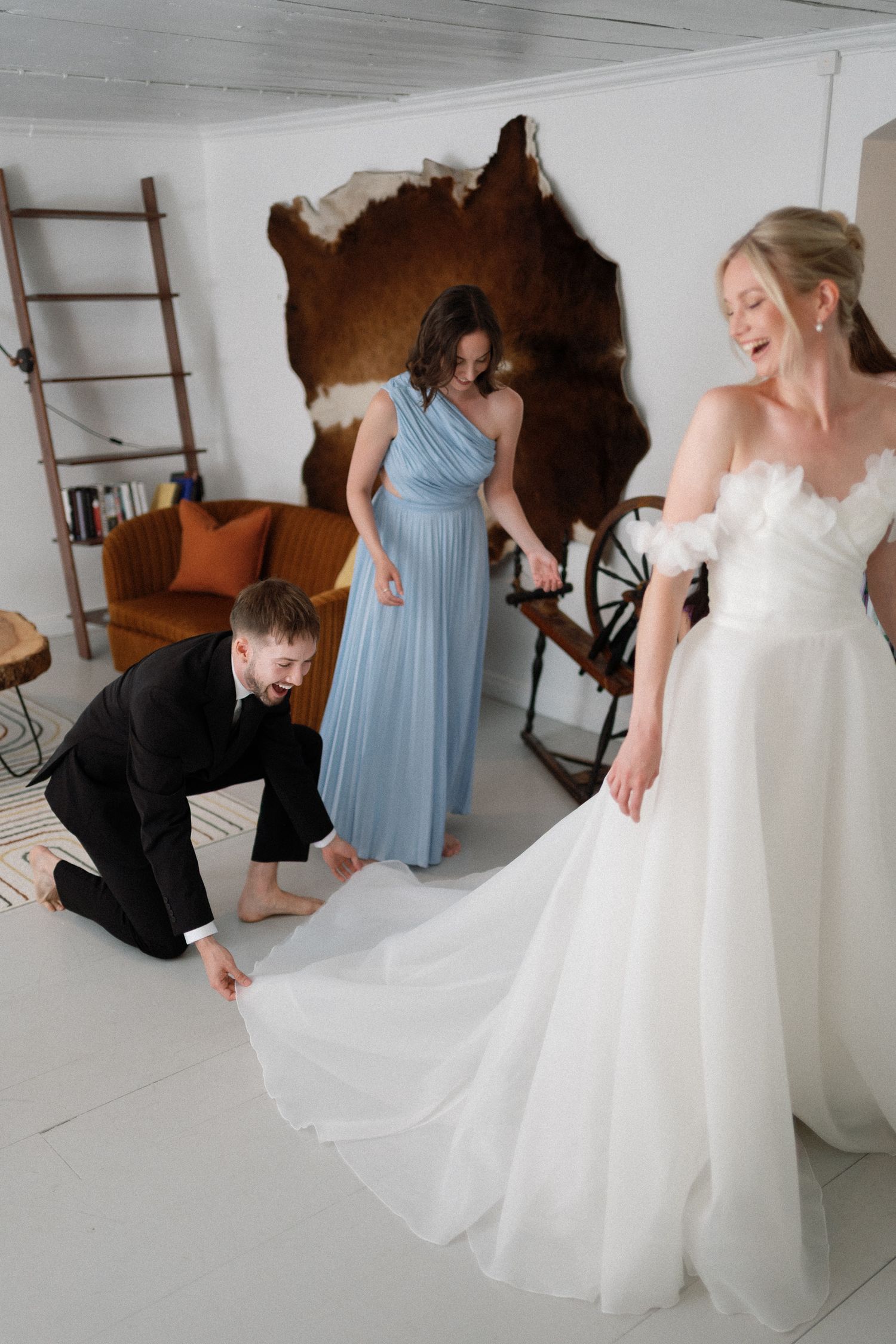 Bride in white gown laughs as bridesmaid in blue dress and groomsman adjust her wedding train indoors.