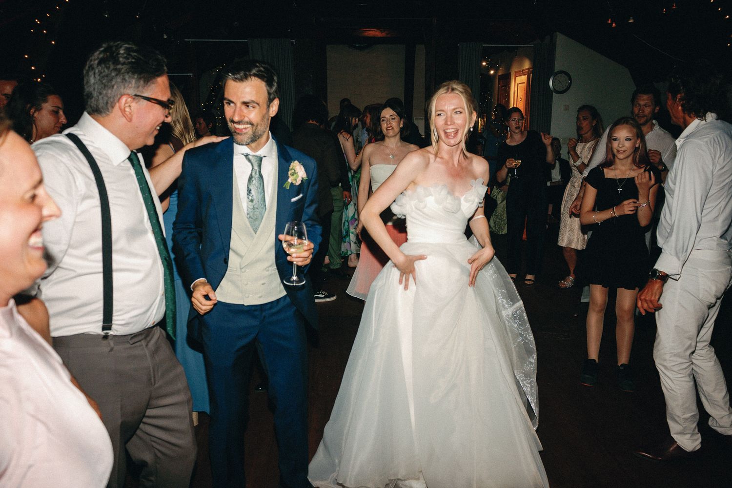Bride in white gown and groom in blue suit dancing joyfully at wedding reception surrounded by guests.