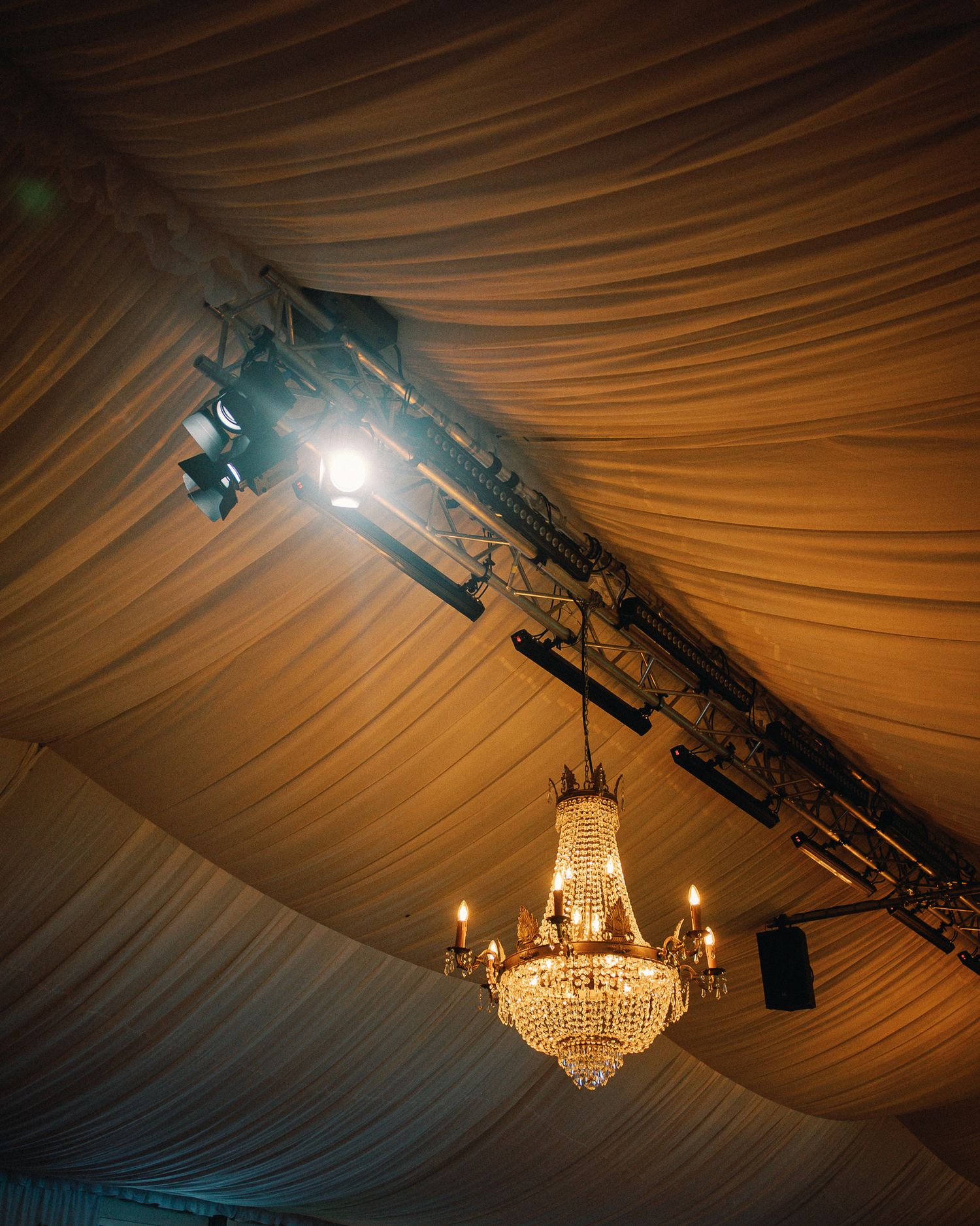 Crystal chandelier and stage lighting hang from draped fabric ceiling in an elegant event tent venue.