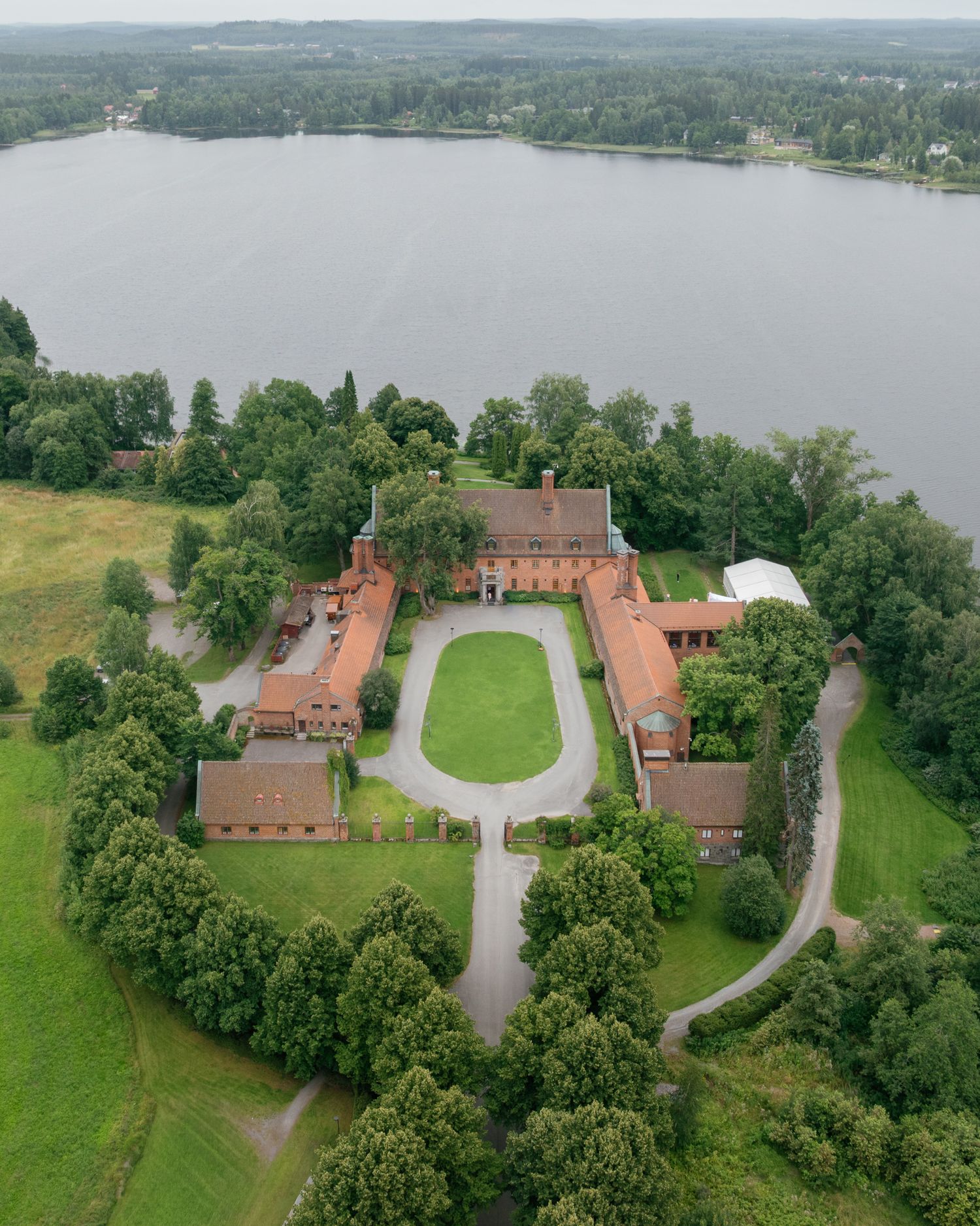 Aerial view of a historic red brick manor estate surrounded by lush trees, manicured grounds, and a serene lake backdrop.