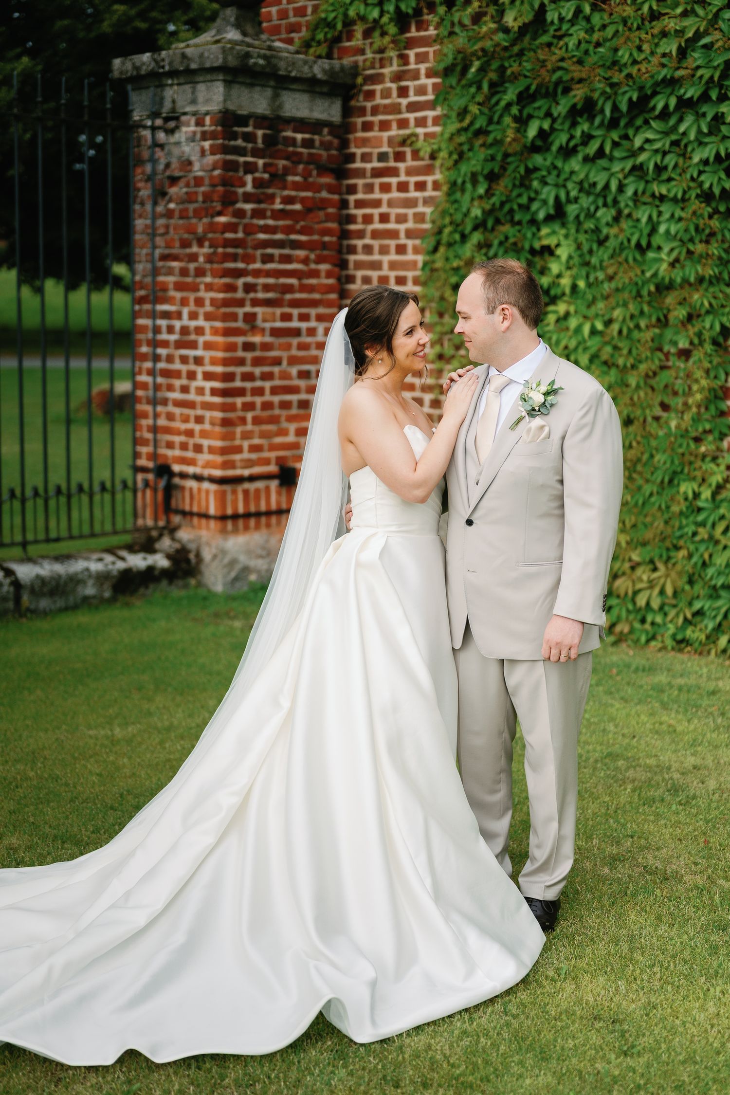 Bride in white ball gown with veil and groom in tan suit pose together on green lawn near brick wall with ivy.