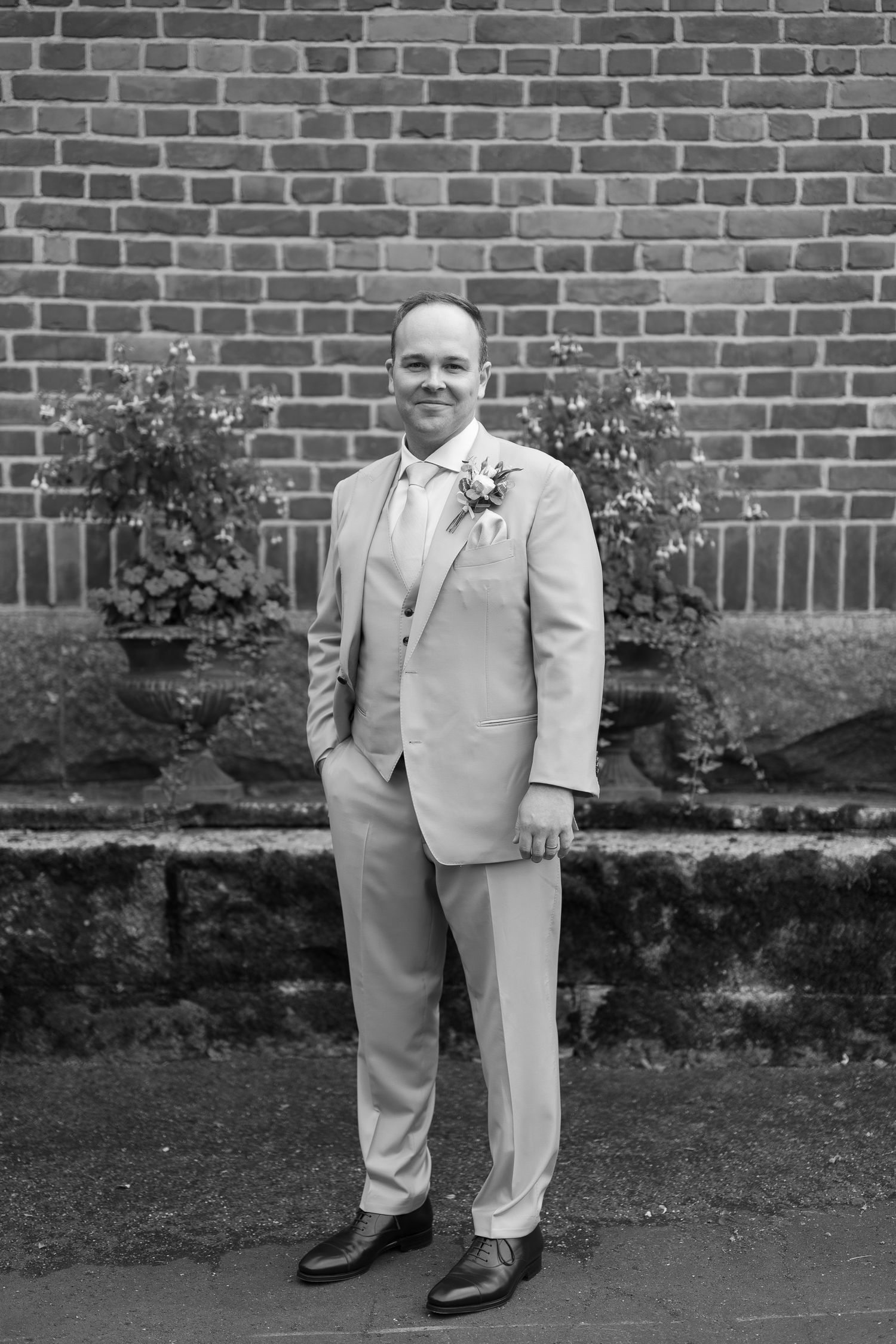 A groom in a light suit with boutonniere poses confidently before a brick wall in this elegant black and white wedding photo.
