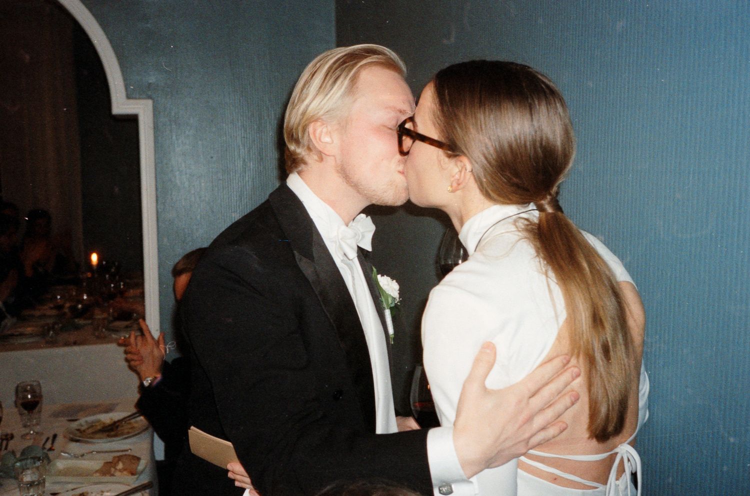 Couple sharing a romantic kiss at a wedding reception, groom in tuxedo and bride in white dress.