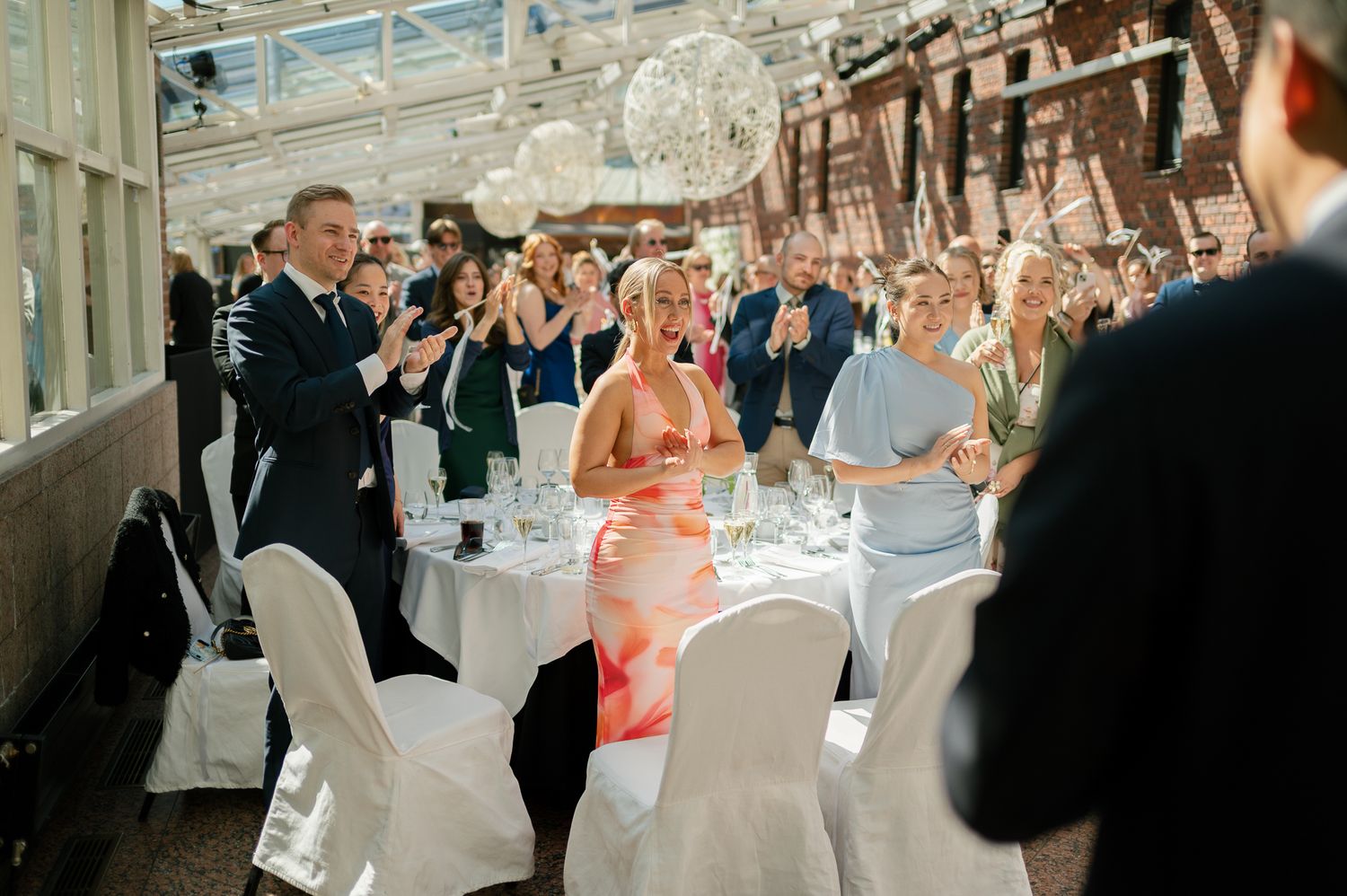 Guests applauding and celebrating at an elegant wedding reception in a bright venue with white chairs and round tables.
