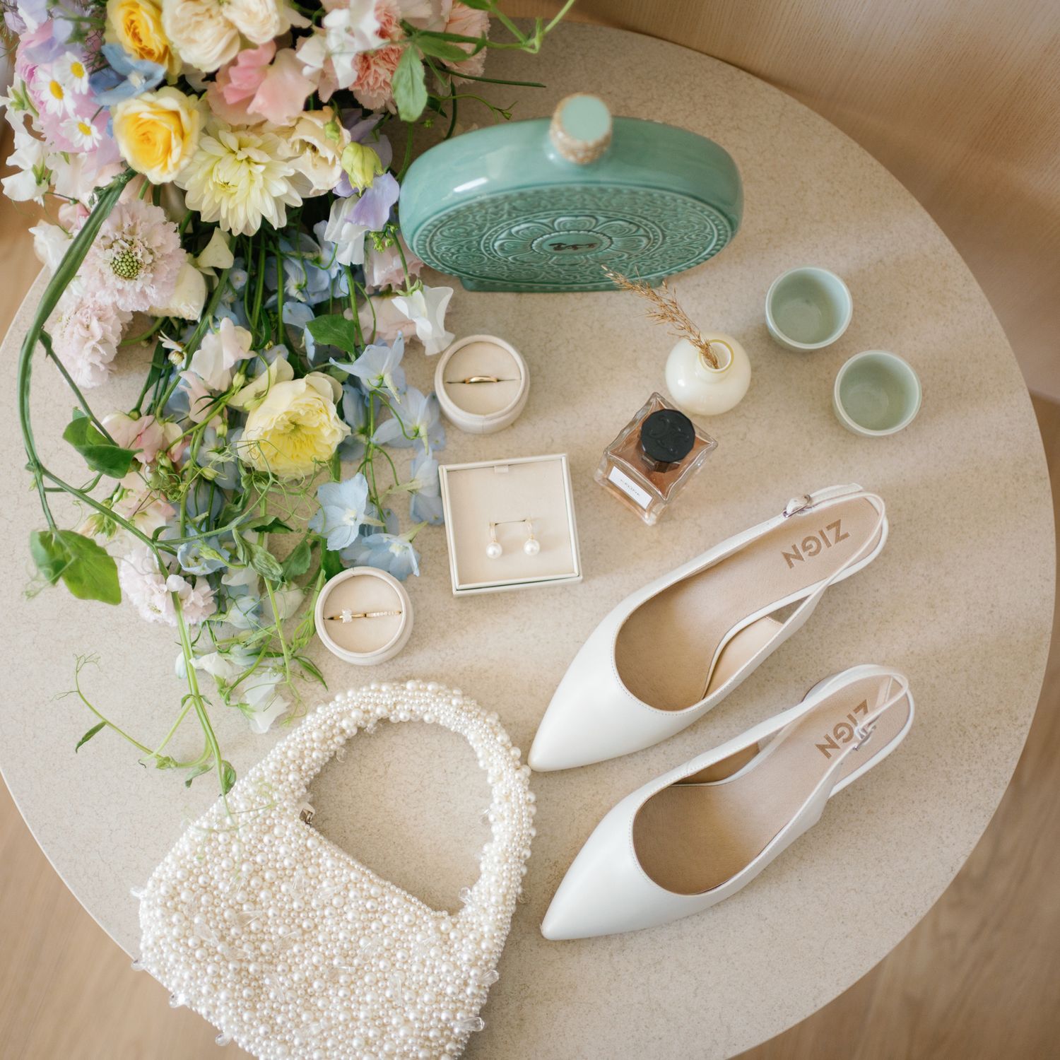 Elegant bridal flat lay with white pearl bag, slingback heels, mint clutch, jewelry boxes, and pastel floral arrangement.