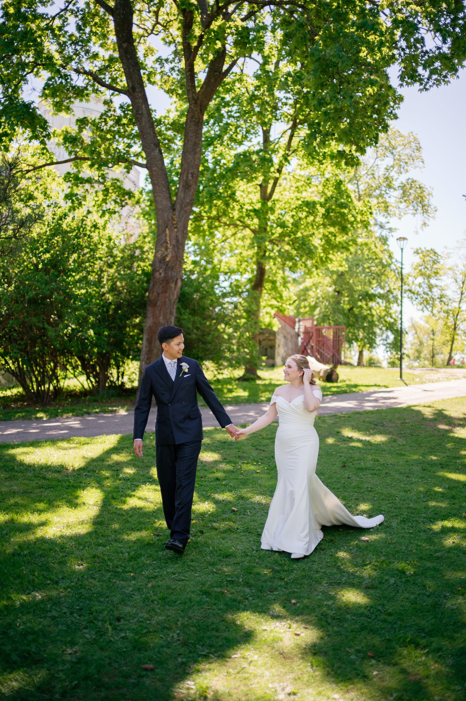 Bride and groom hold hands walking through a sunlit park, bride in white mermaid gown, groom in dark suit.