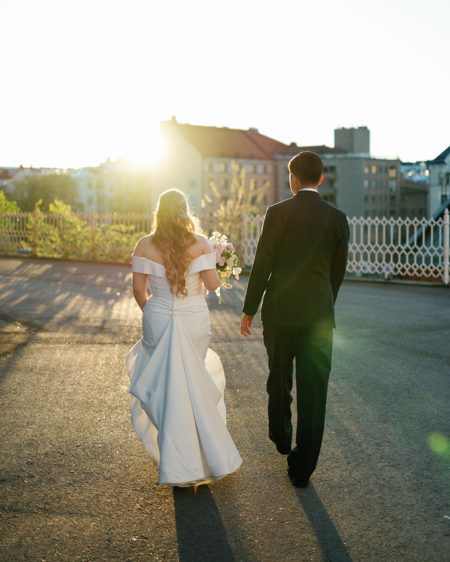 Bride and groom walking hand in hand at sunset, bride in off-shoulder white gown holding bouquet, groom in black suit.