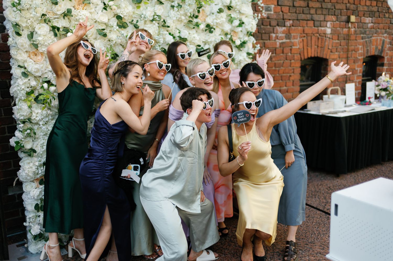 A joyful group of friends wearing heart sunglasses celebrate at a wedding reception near a floral wall.