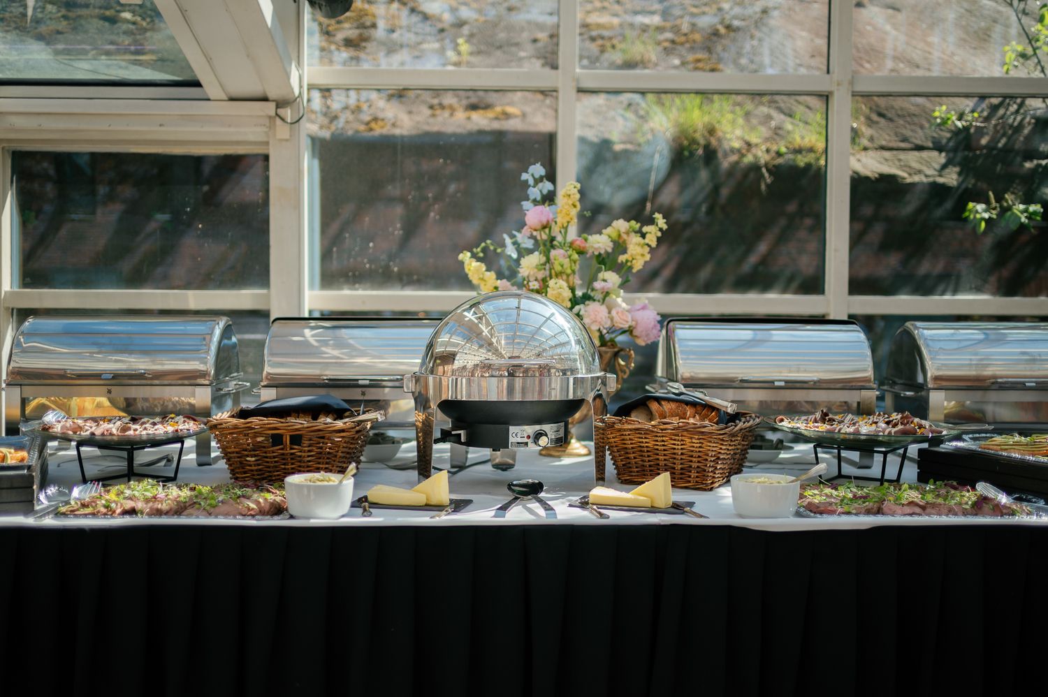 Elegant buffet table with wicker baskets, chafing dishes, and floral centerpiece set near large windows.