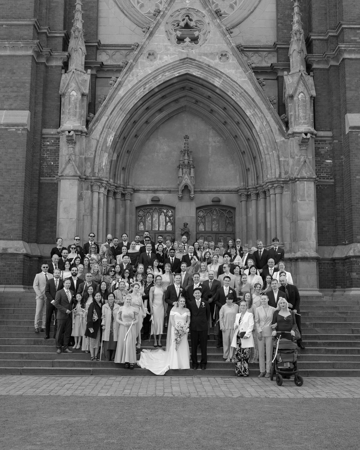 Black and white wedding photo of a large group posing on church steps beneath a Gothic stone archway.