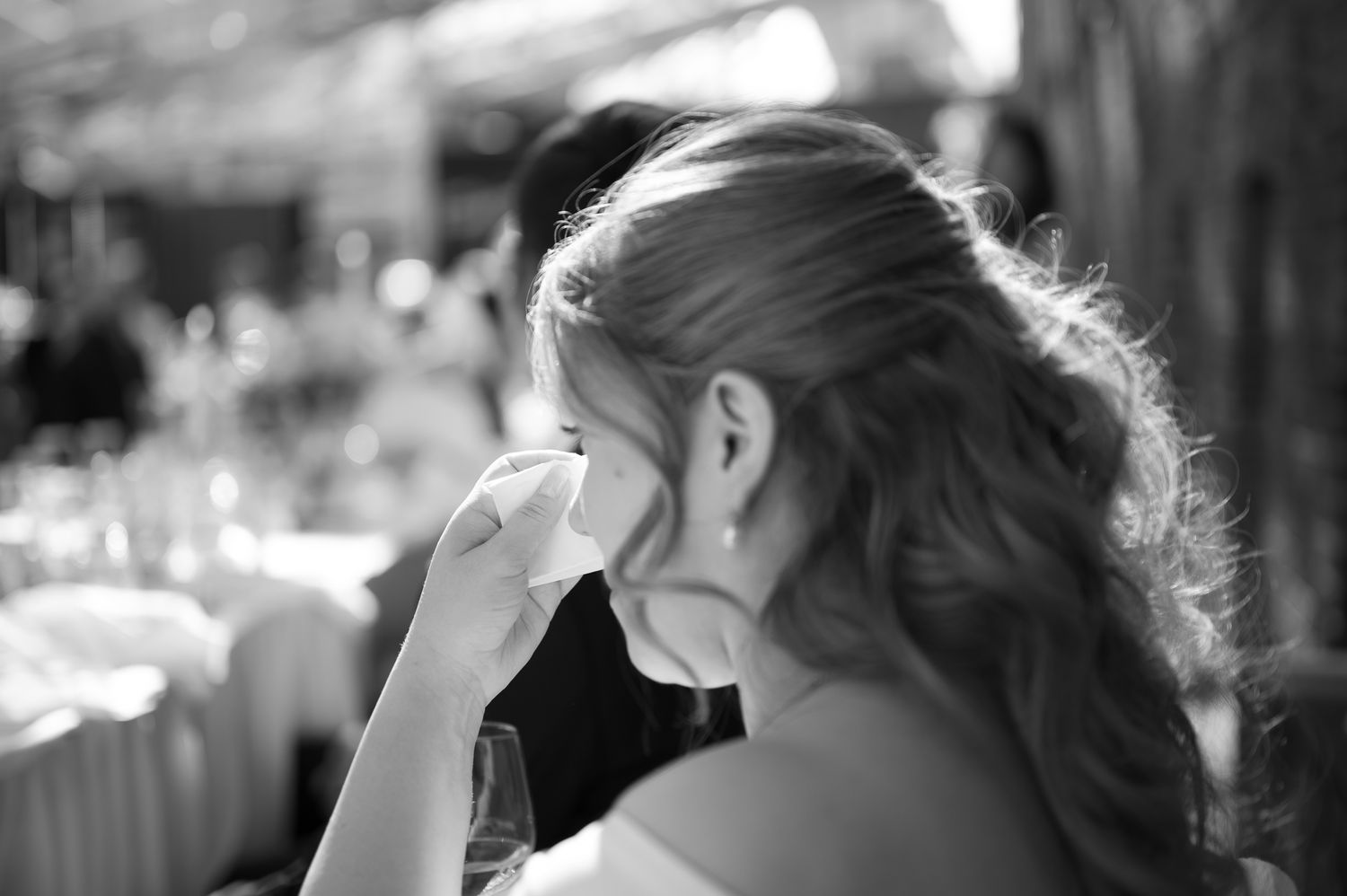 Black and white photo of a woman wiping tears at a wedding reception, captured from behind.