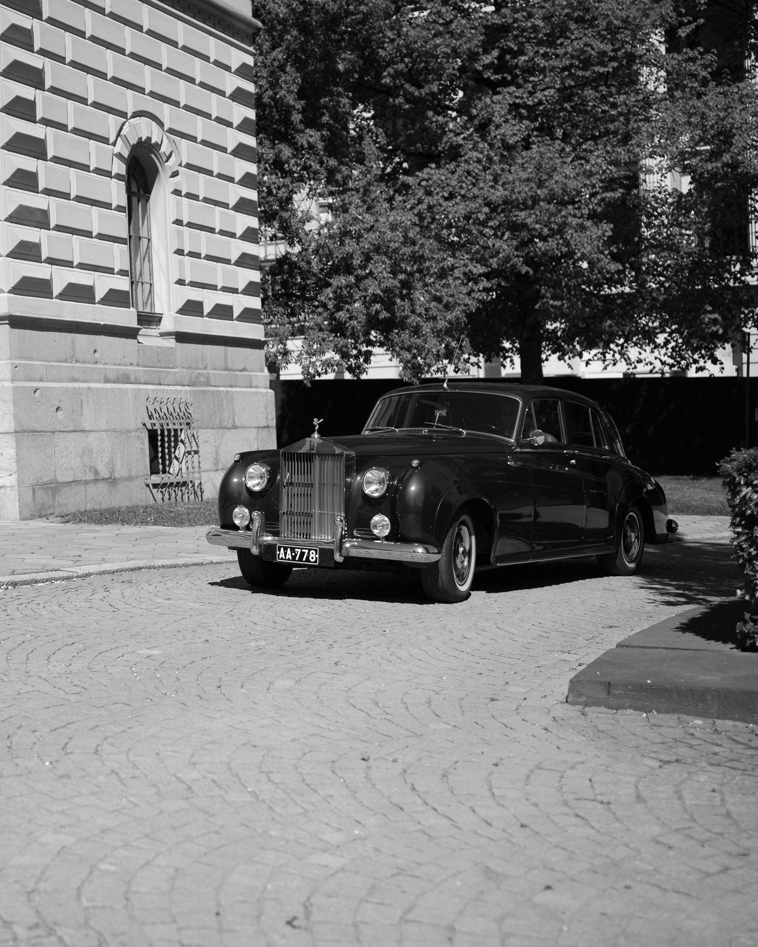 Vintage black Rolls-Royce parked on cobblestone driveway beside classical building with trees in background, black and white photo.