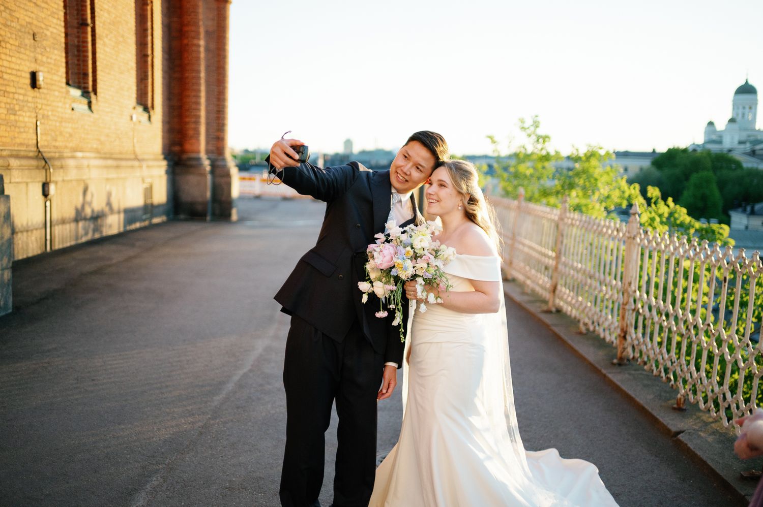 Bride and groom taking a selfie on a sunny day, bride holding a bouquet in a white gown, groom in a black suit.