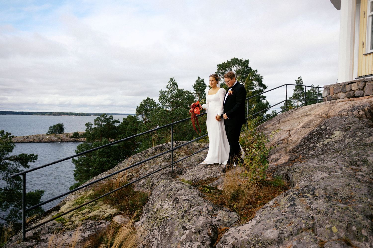 A couple stands on a rocky cliff overlooking a scenic waterfront landscape at sunset.