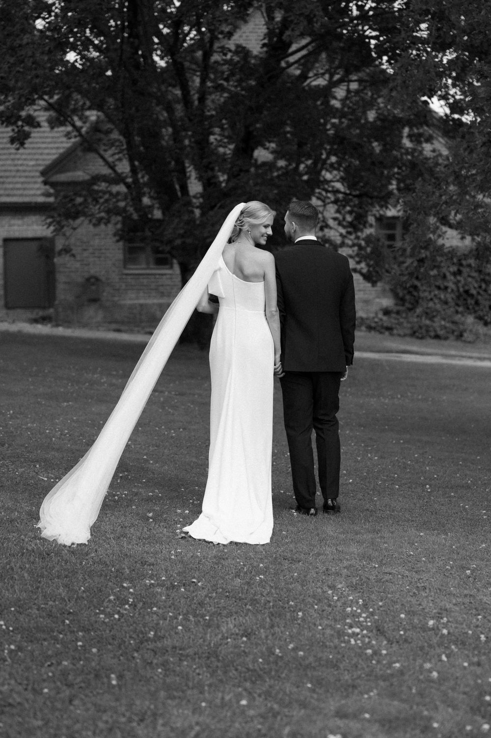 A flowing white wedding dress trails behind as a couple walks across a garden lawn.