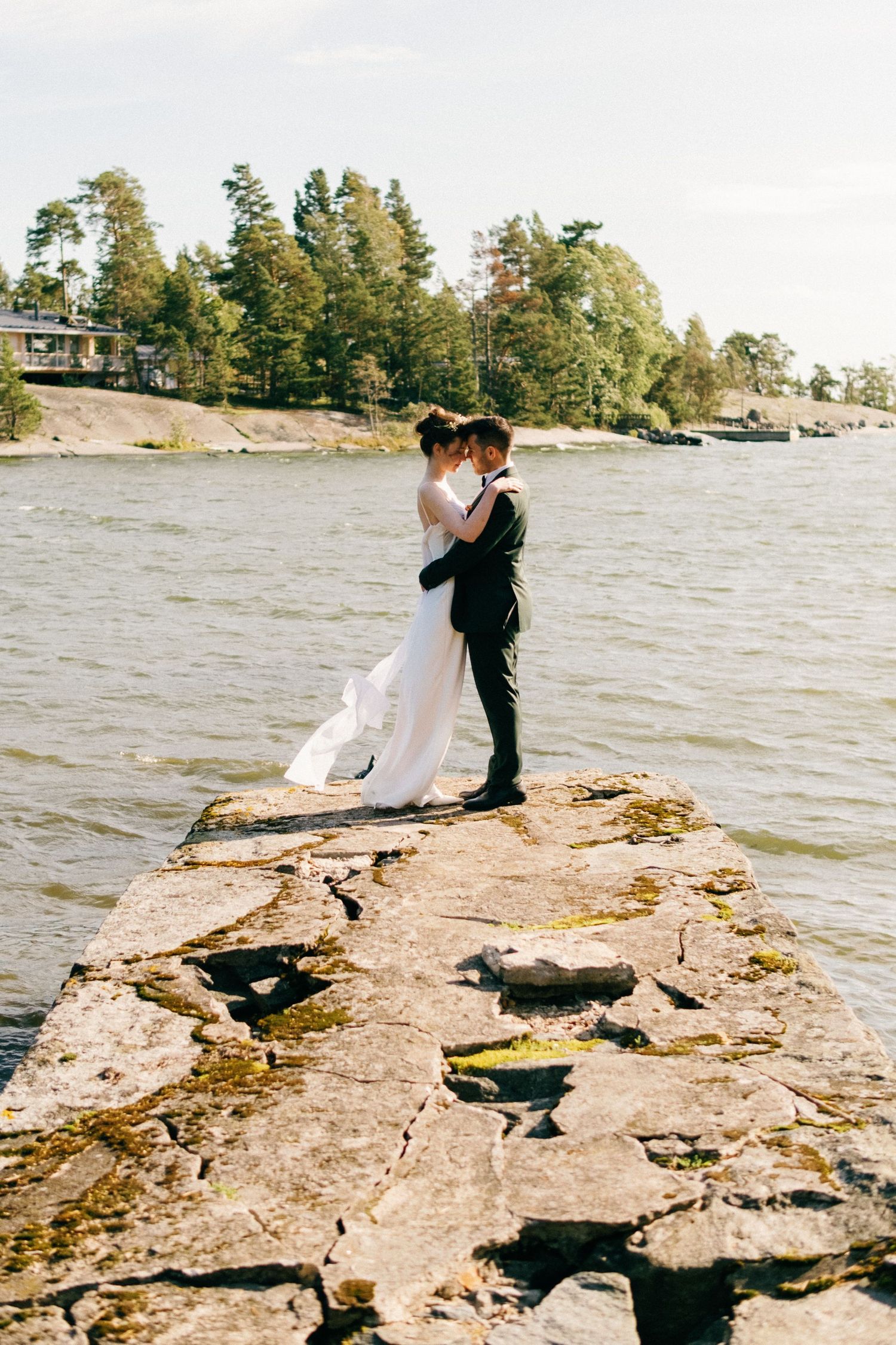 A couple sits on a rocky shoreline overlooking a lake surrounded by trees on a sunny day.