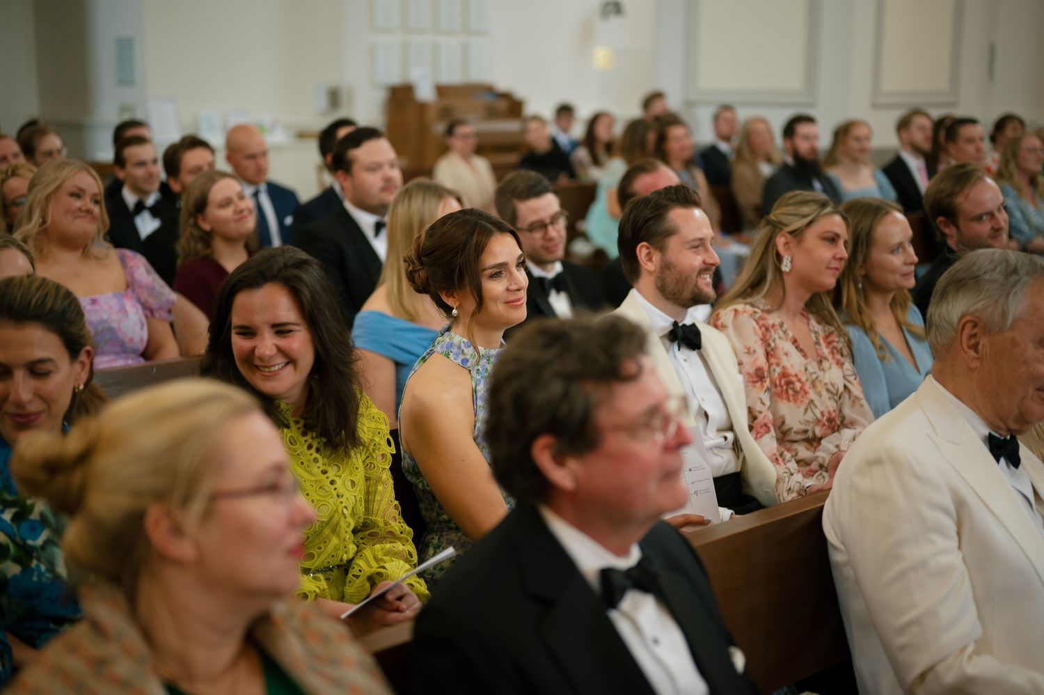 Wedding guests react with joy and laughter while watching a ceremony from their seats.