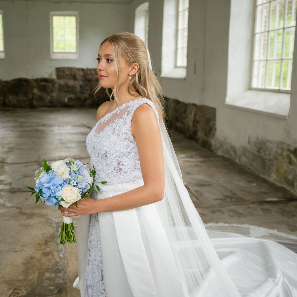 A bride in a white lace gown holds a blue bouquet in a sunlit room with vintage character.
