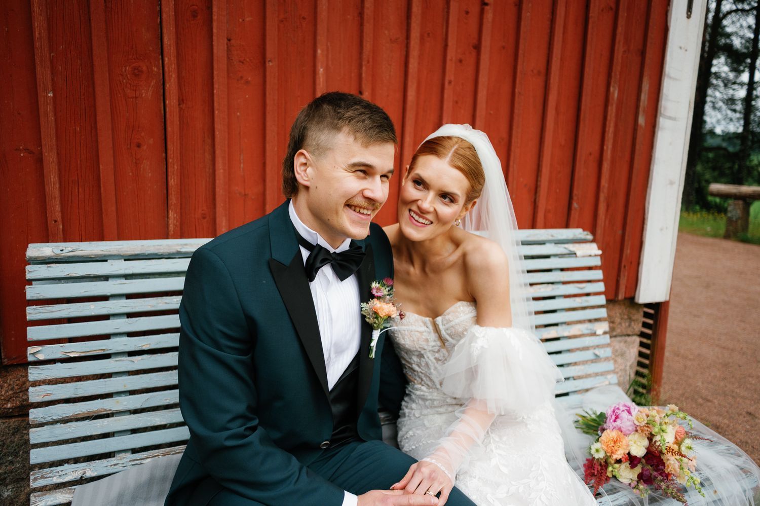 Newlyweds pose together on white bench against rustic red barn backdrop.