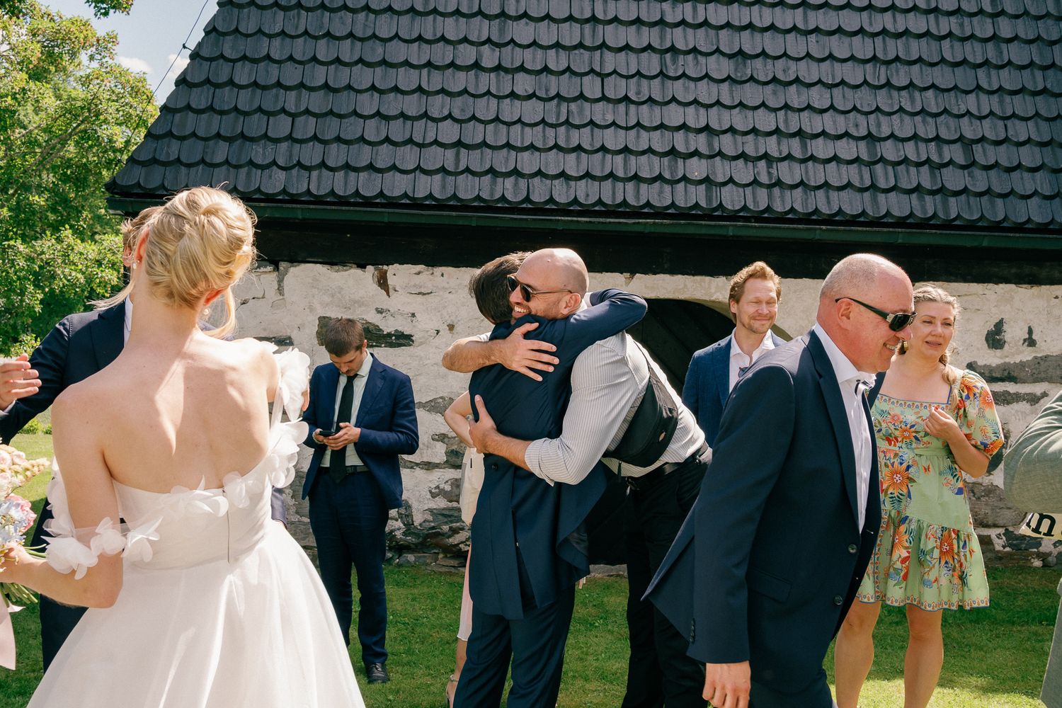 Groom hugging a guest at an outdoor wedding reception near a rustic stone building on a sunny day.