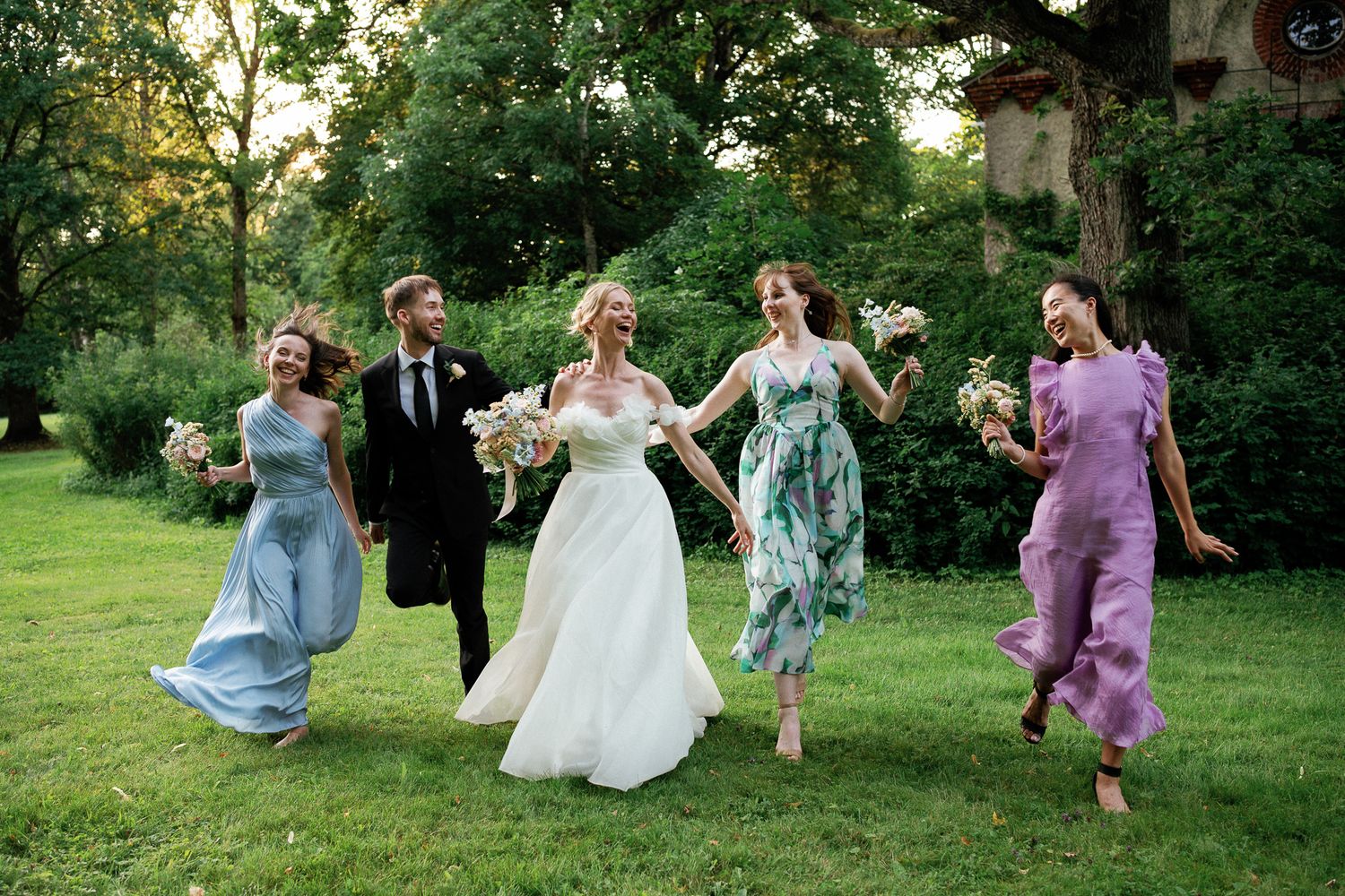 A joyful wedding party runs across green grass, with bride in white gown and attendants in colorful dresses holding bouquets.