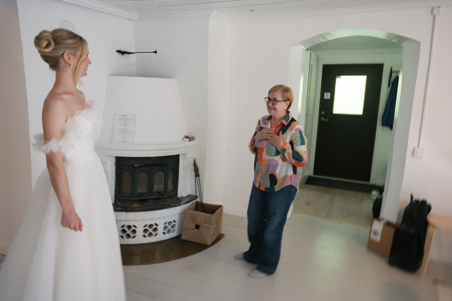 A bride in a white off-shoulder wedding dress stands near a fireplace while a woman in casual attire photographs her indoors.