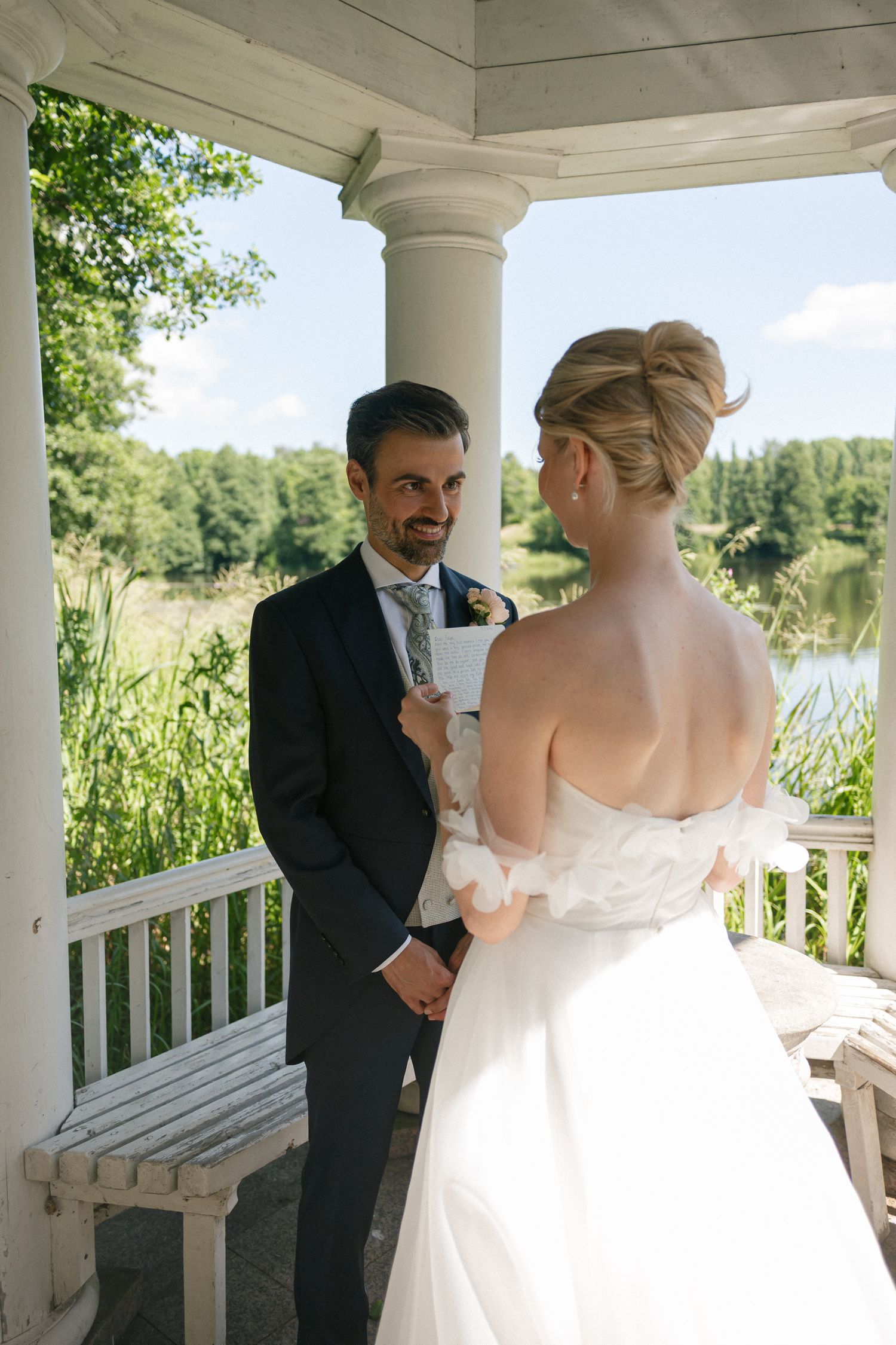 Bride and groom share a romantic moment in a white gazebo overlooking a serene lake surrounded by lush greenery.