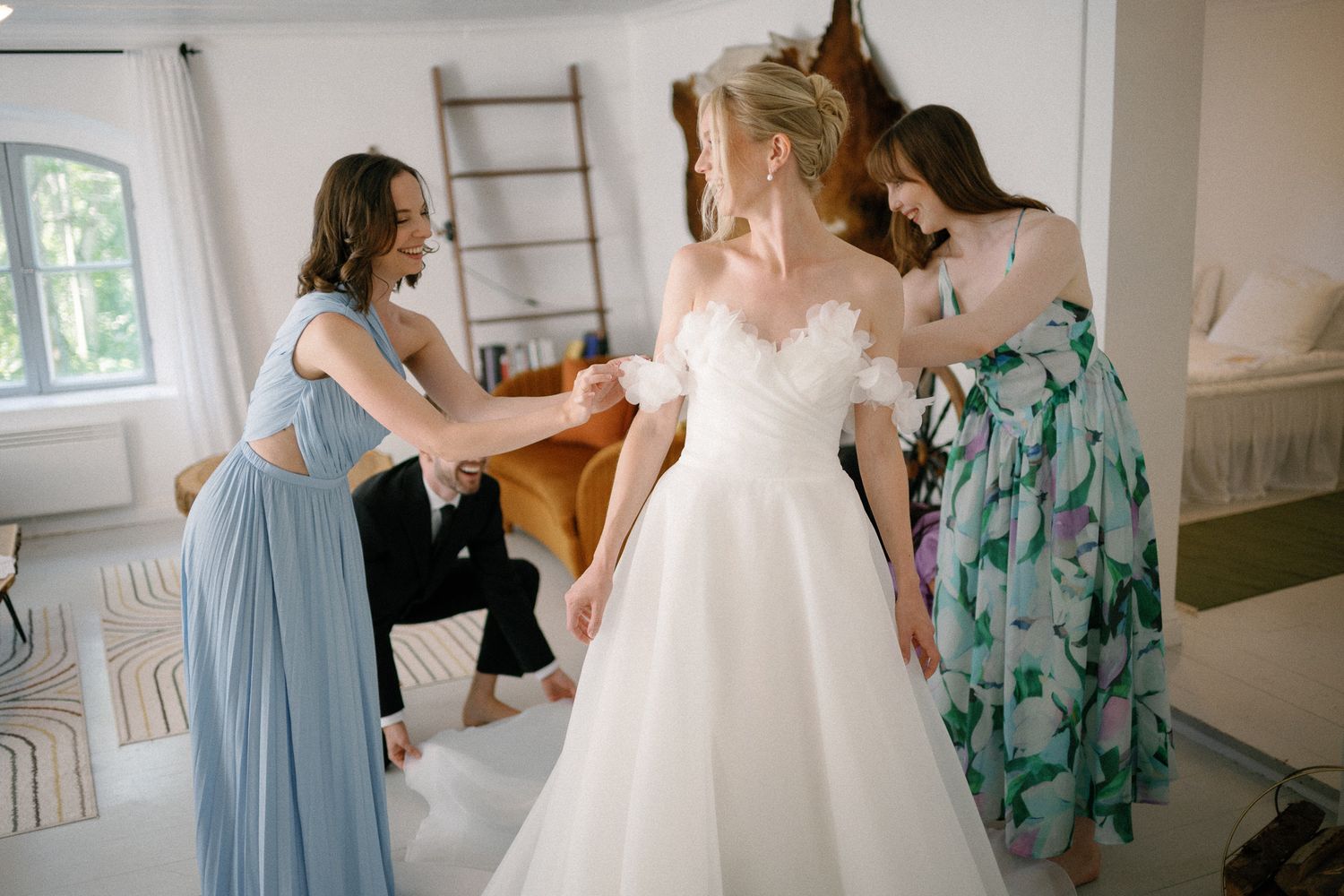 Bridesmaids helping bride into white wedding gown in bright white room before ceremony.