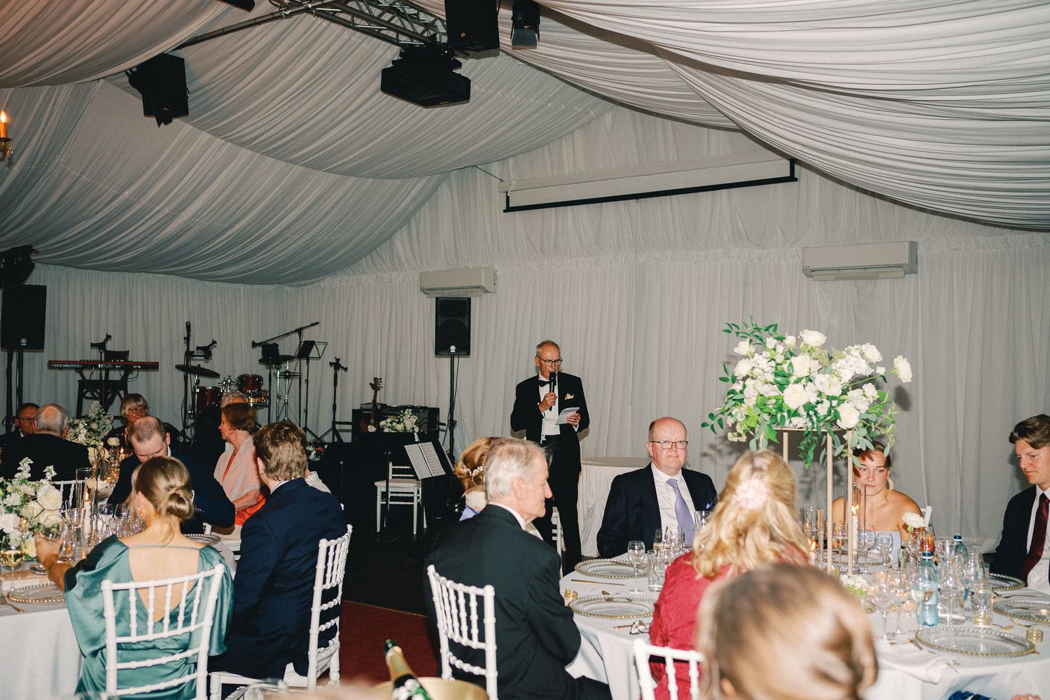Elegant wedding reception in a draped marquee with guests seated at decorated tables while a man delivers a speech.