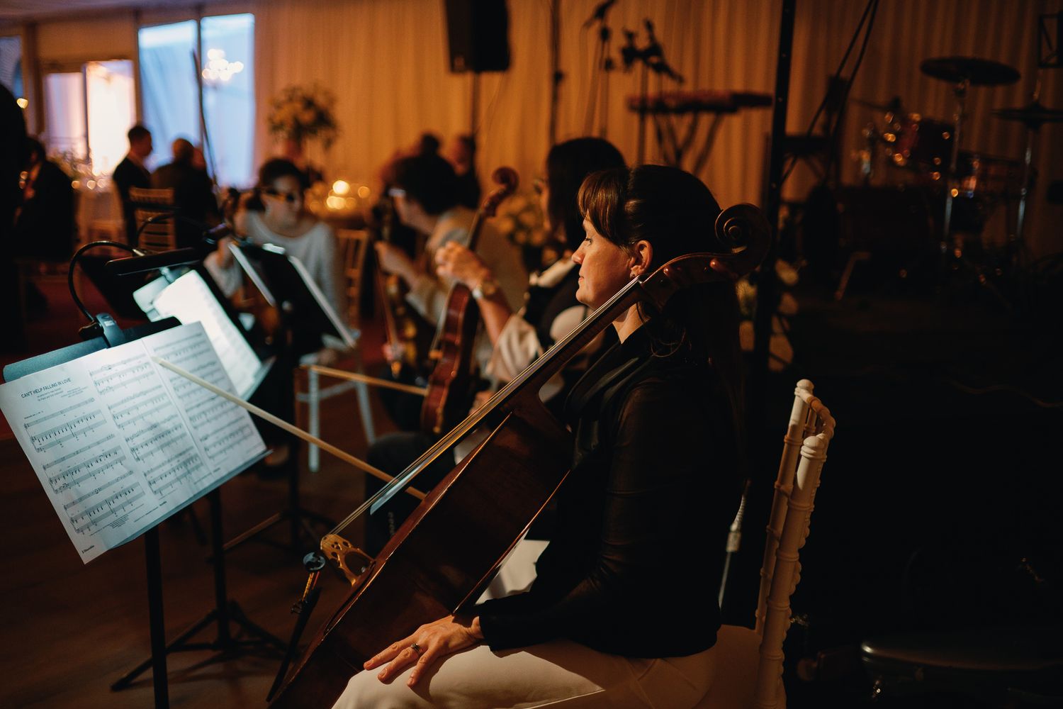 A cellist performs sheet music at an elegant candlelit event with orchestra musicians in the background.