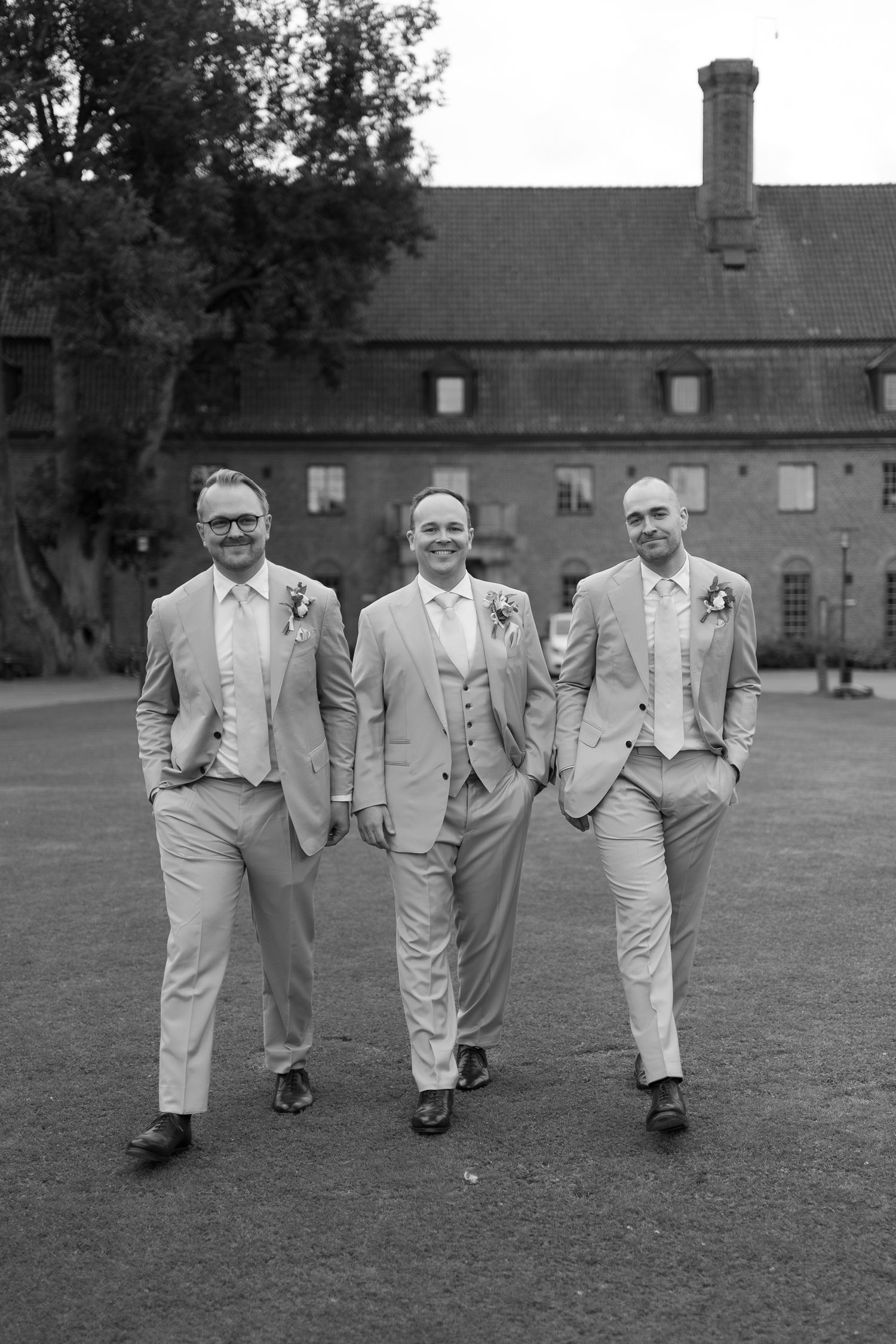 Three groomsmen in matching light suits with boutonnieres pose outdoors before a brick building in black and white photo.