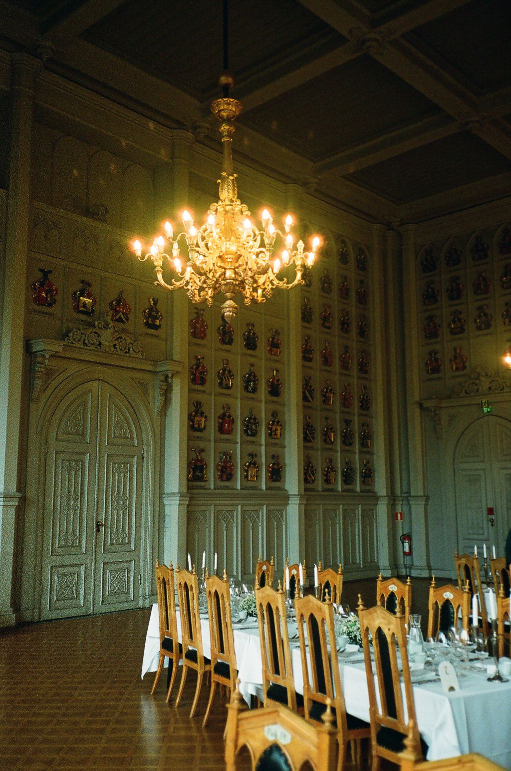 Elegant banquet hall with golden chandelier, long dining table, candles, and heraldic coat of arms panels on walls.