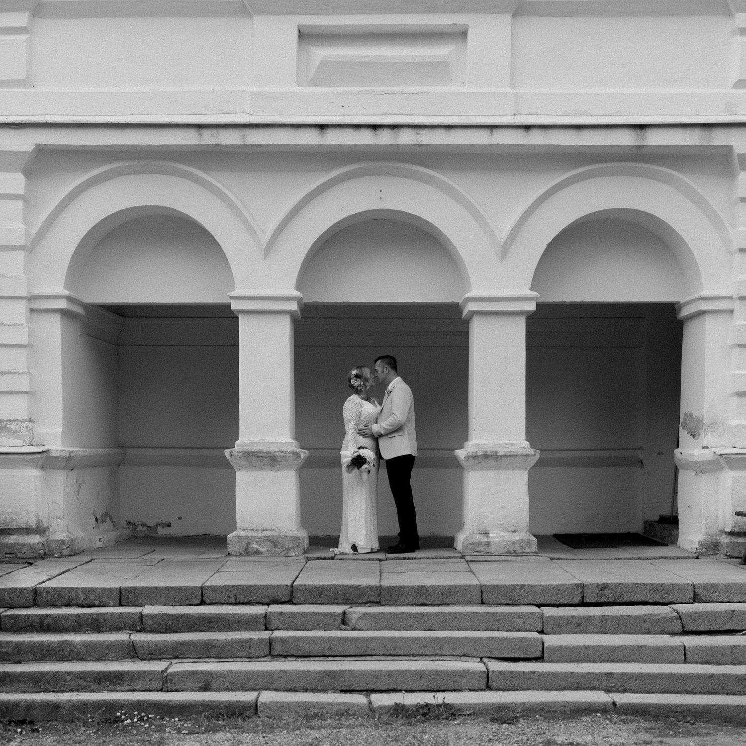 Black and white photo of a couple embracing under elegant arched columns of a classical building.
