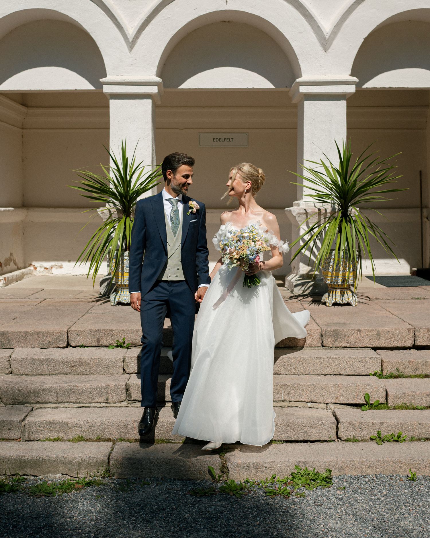 Bride and groom walk hand in hand on stone steps before arched building with tropical plants on sunny wedding day.