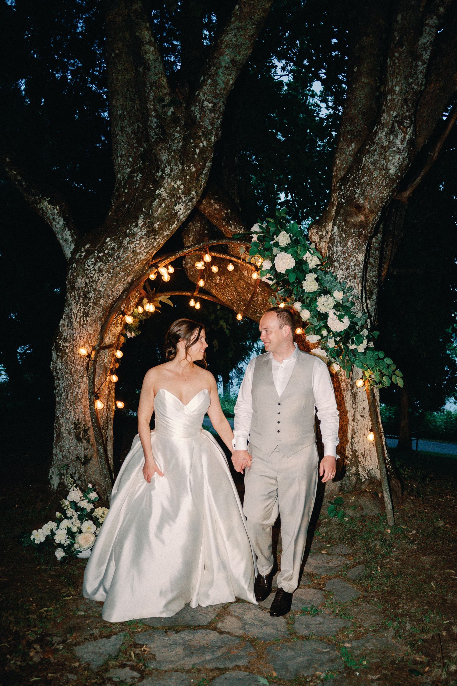 Bride and groom hold hands under a floral arch with string lights between trees at an outdoor evening wedding.