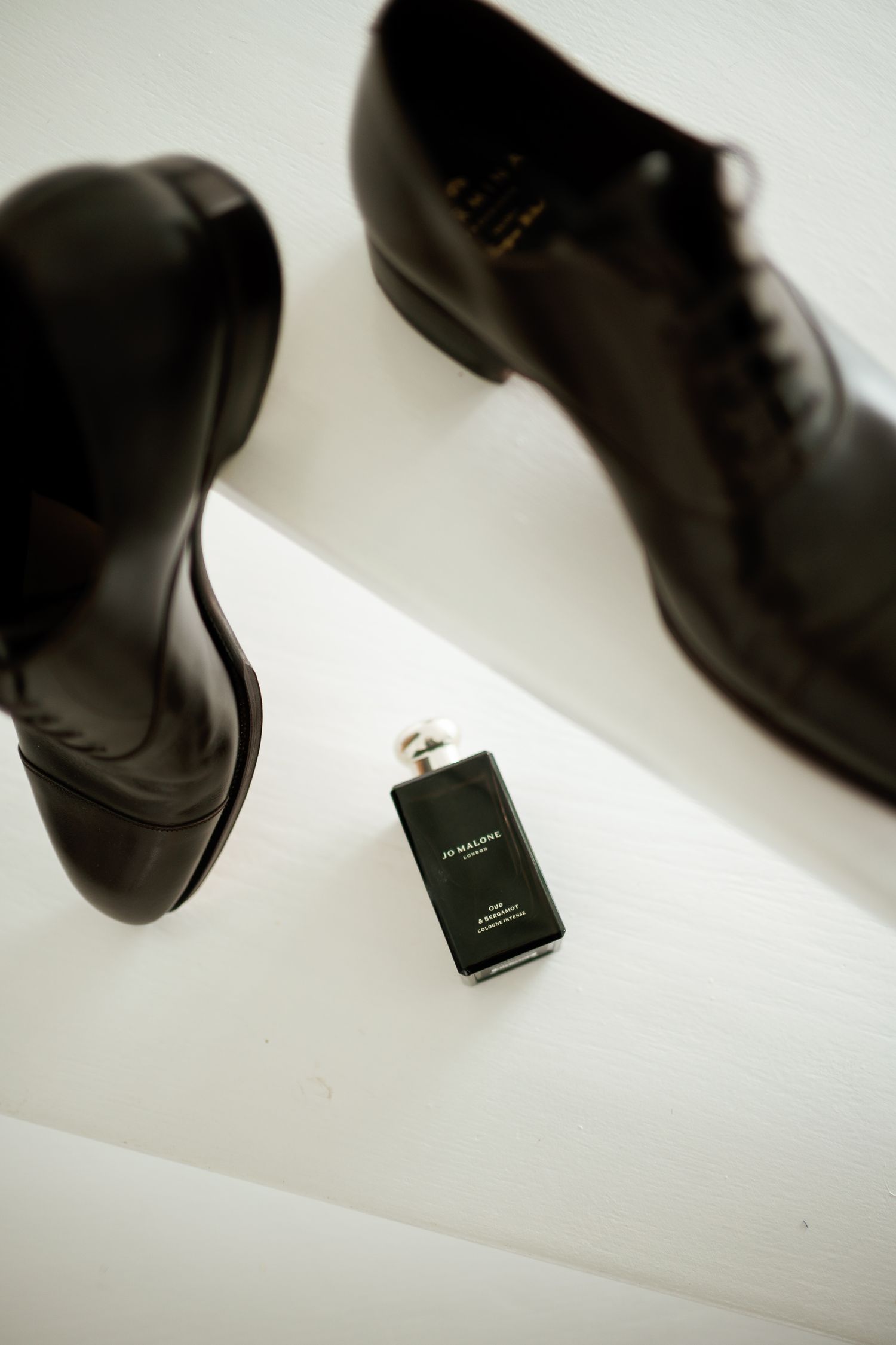 Black leather Oxford dress shoes beside a Jo Malone cologne bottle on a white surface.