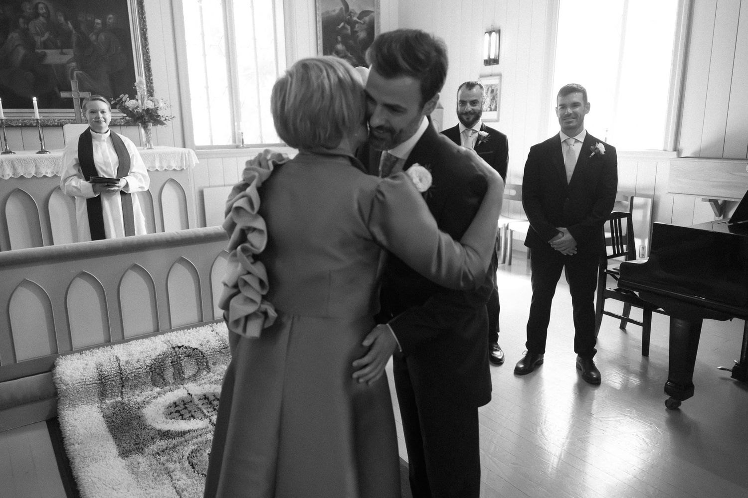 Black and white wedding photo of groom embracing a guest inside a church while others look on.