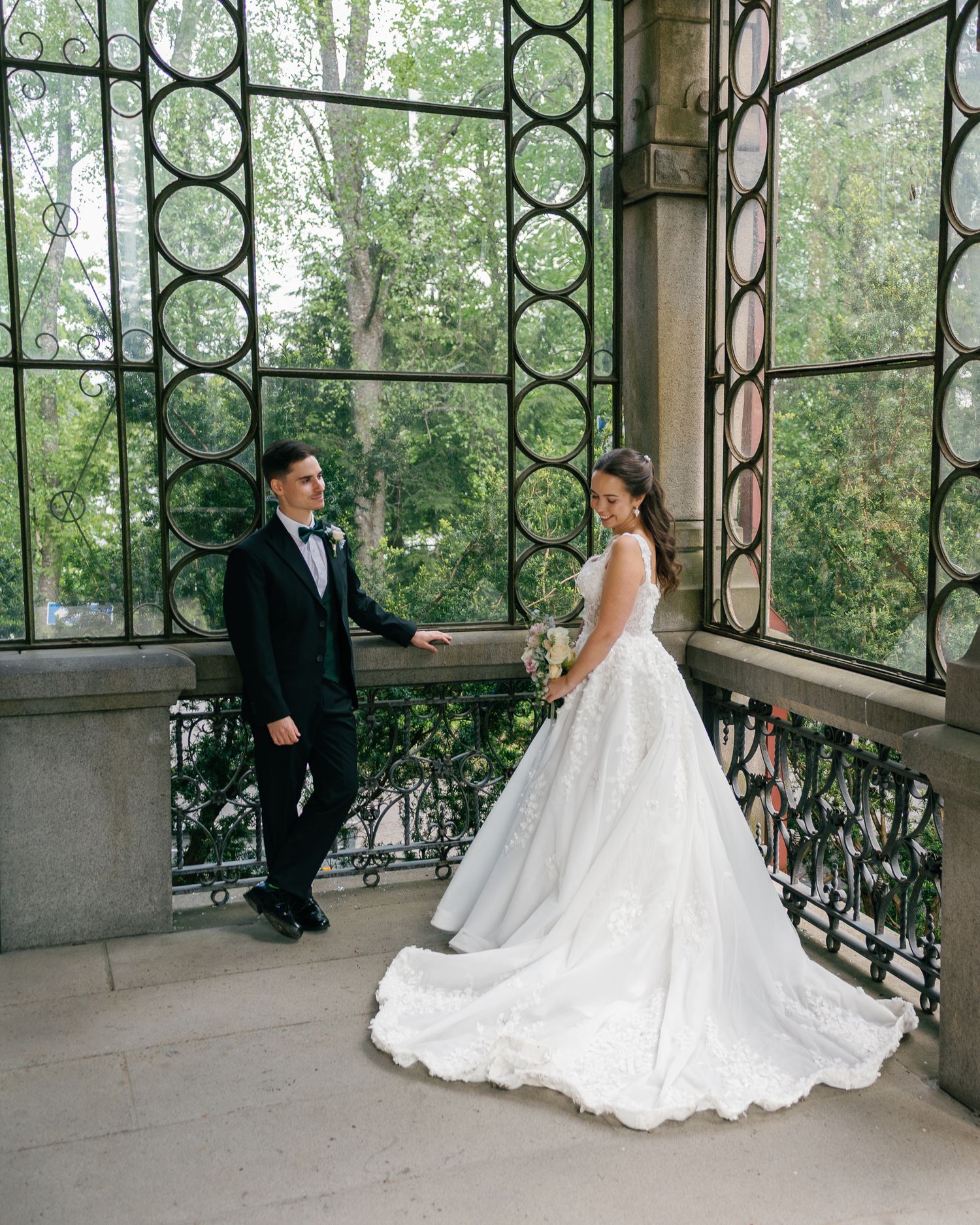 Bride in white ball gown and groom in black tuxedo pose on ornate balcony with decorative iron lattice windows.