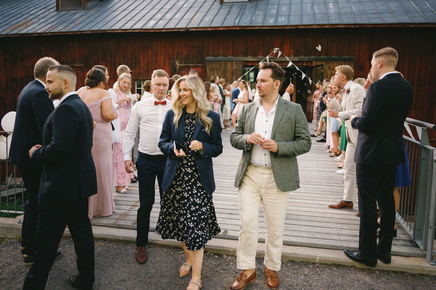 Wedding guests mingle on a rustic wooden deck outside a barn during a casual reception celebration.
