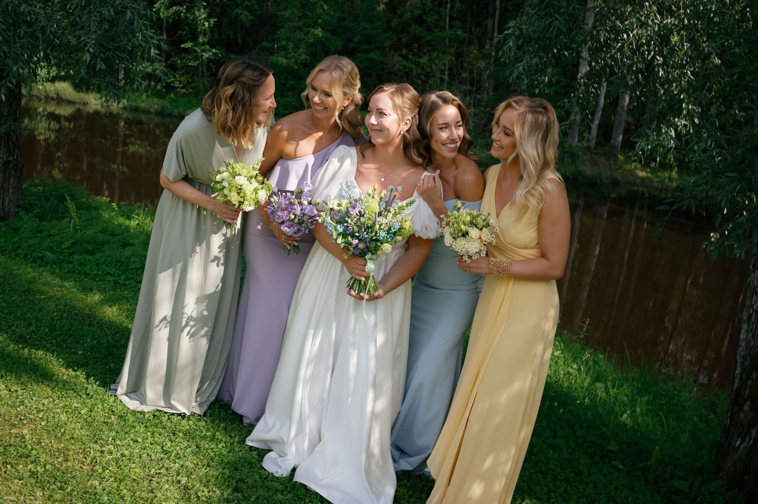 Group photo of wedding party members in pastel dresses holding floral bouquets in an outdoor setting.