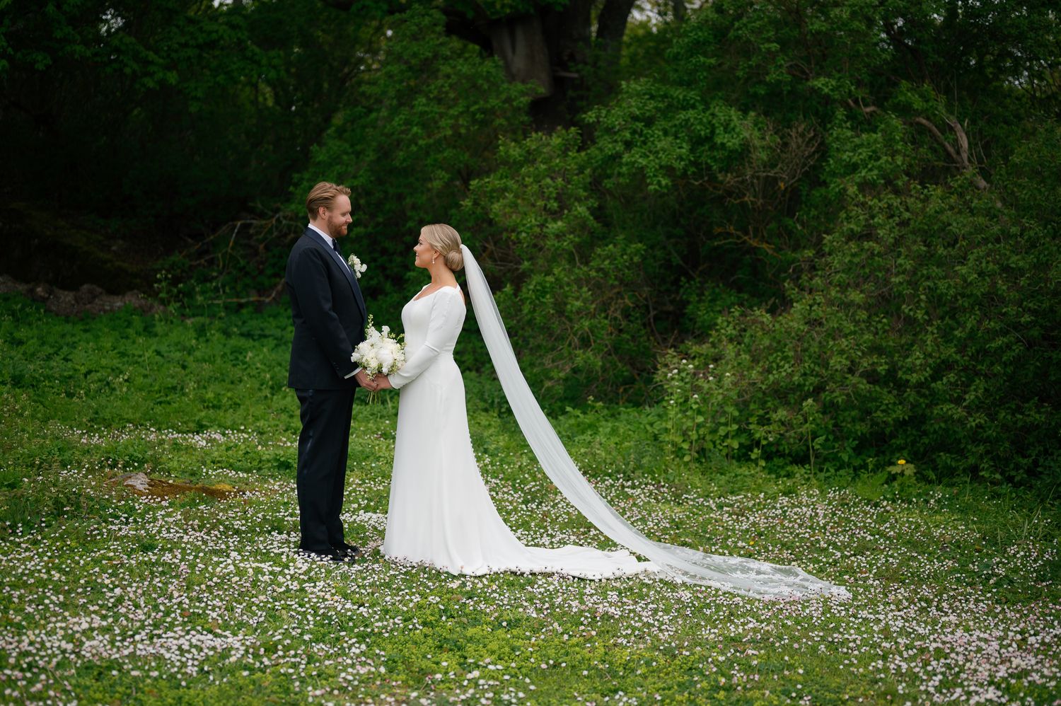 A couple in wedding attire stands together in a lush green garden setting with scattered white flowers on the ground.
