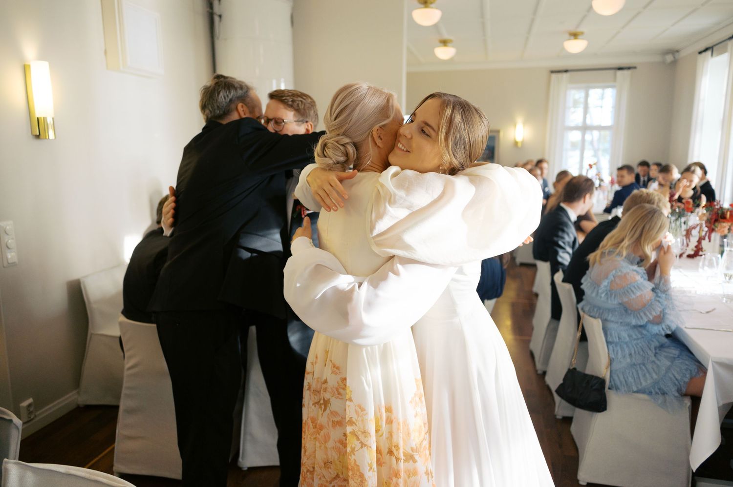 Wedding guests embrace in a heartfelt group hug during an indoor reception celebration.