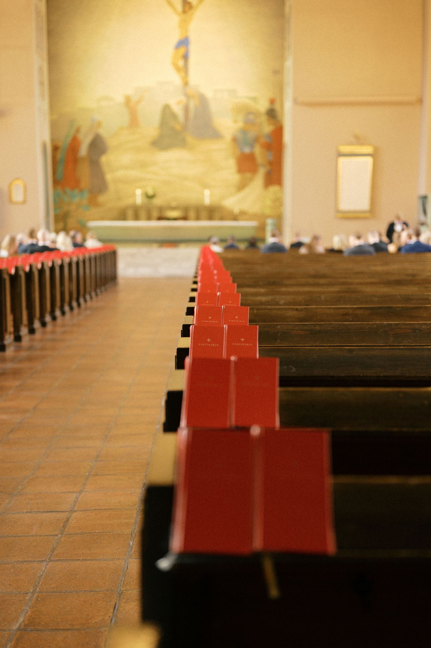 Rows of wooden church pews with red prayer book holders line up in a traditional sanctuary with tiled floors.