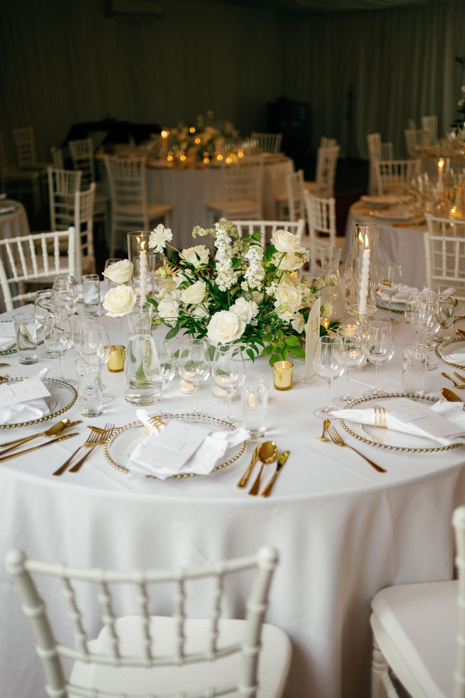 Elegant wedding reception table with white linen, gold cutlery, crystal glasses, and white floral centerpiece.