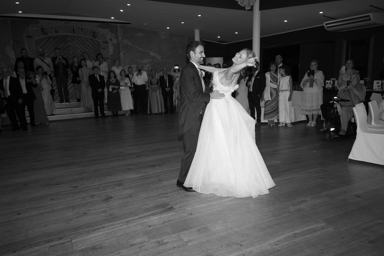 Black and white photo of a bride and groom sharing their first dance at a wedding reception with guests watching.