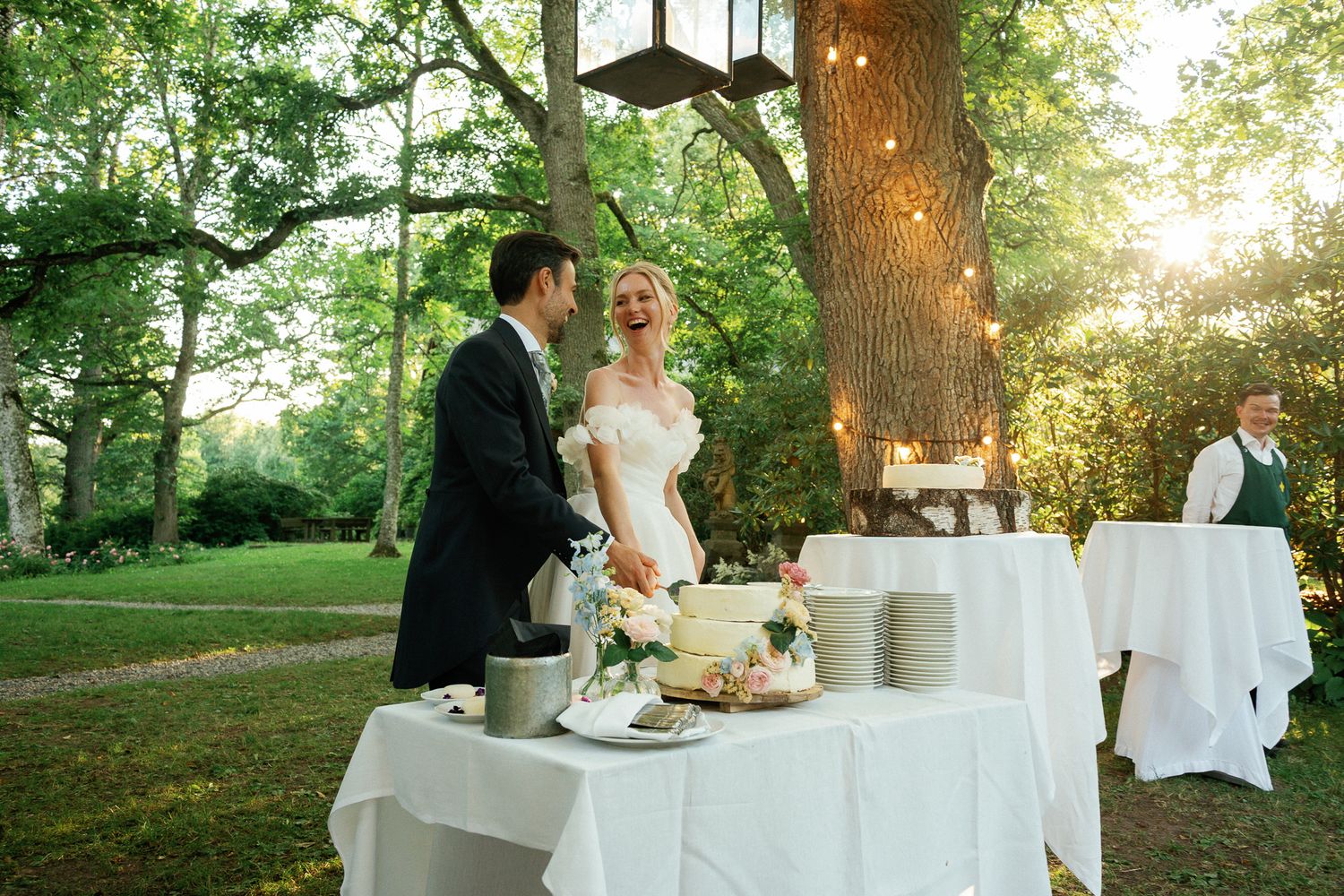 Bride and groom cutting wedding cake outdoors under trees with string lights at elegant garden reception.