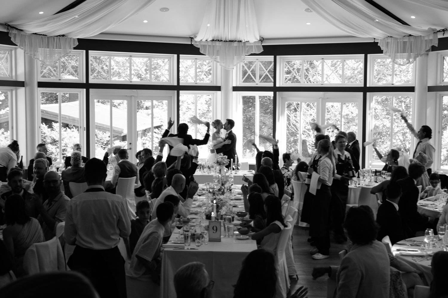 Black and white photo of a lively wedding reception with guests gathered around tables in an elegant windowed venue.